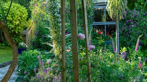 A lush garden with various plants and flowers, a wooden pergola, and a glass structure in the background.