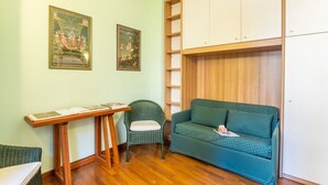 Une pièce avec un canapé vert, un fauteuil en osier, une table en bois sur laquelle sont posés des livres, et deux tableaux encadrés accrochés au mur.
