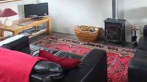 A living room with a black leather sofa, a red throw blanket, a television on a wooden table, a fireplace, and a bookshelf. - Grooms Cottage- Sleeps 4 in 2 bedrooms plus 2 to 4 dogs depending on size. (Bodmin)