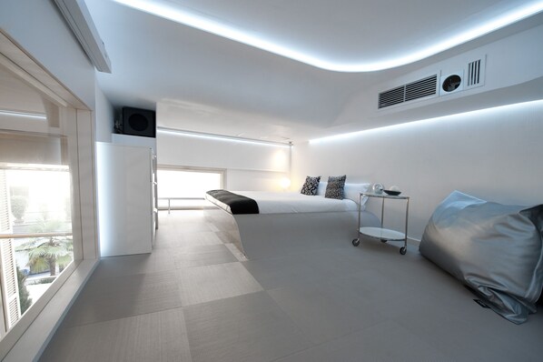 A modern hotel room with a large bed, a bedside table, a window with a view, and a ceiling with recessed lighting.