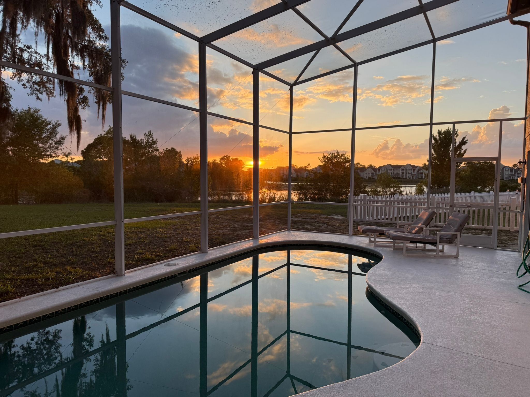 A swimming pool with a glass enclosure, surrounded by a deck with lounge chairs and a view of a sunset over a residential area.
