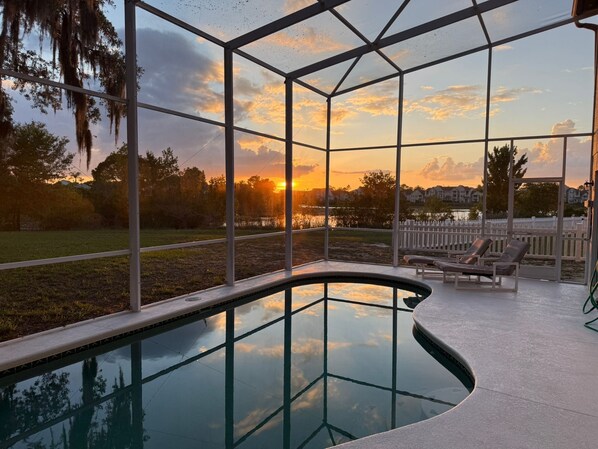 A swimming pool with a glass enclosure, surrounded by a deck with lounge chairs and a view of a sunset over a residential area.