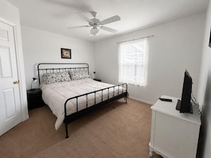 A bedroom with a bed, a ceiling fan, a window with blinds, a nightstand, and a television on a stand.