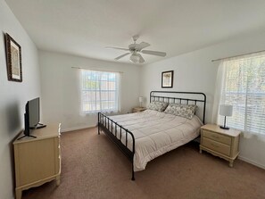 A bedroom with a bed, a ceiling fan, a TV, a bedside table, a nightstand, and a window with blinds.