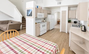 A compact kitchen with white appliances, a dining area with a checkered tablecloth, and a staircase leading to another level.