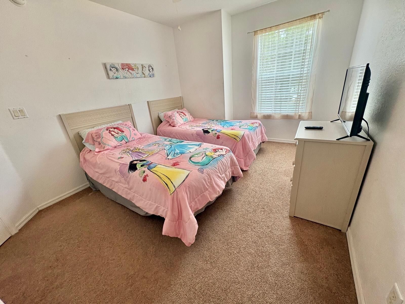 Une chambre avec deux lits, chacun recouvert d’une couverture rose ornée de personnages de dessins animés, un bureau avec un ordinateur et une fenêtre avec des stores.