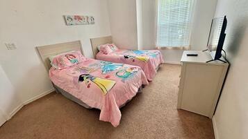 Une chambre avec deux lits, chacun recouvert d’une couverture rose ornée de personnages de dessins animés, un bureau avec un ordinateur et une fenêtre avec des stores.