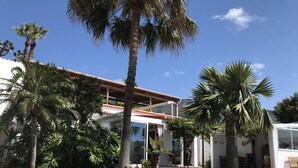 Ein zweistöckiges Haus mit rotem Ziegeldach, umgeben von üppiger Vegetation und Palmen.