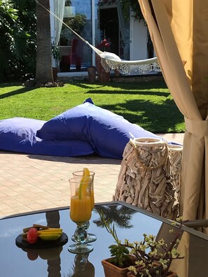A glass table with a drink, fruit, and a small plant. There is a hammock and a cushion in the background.