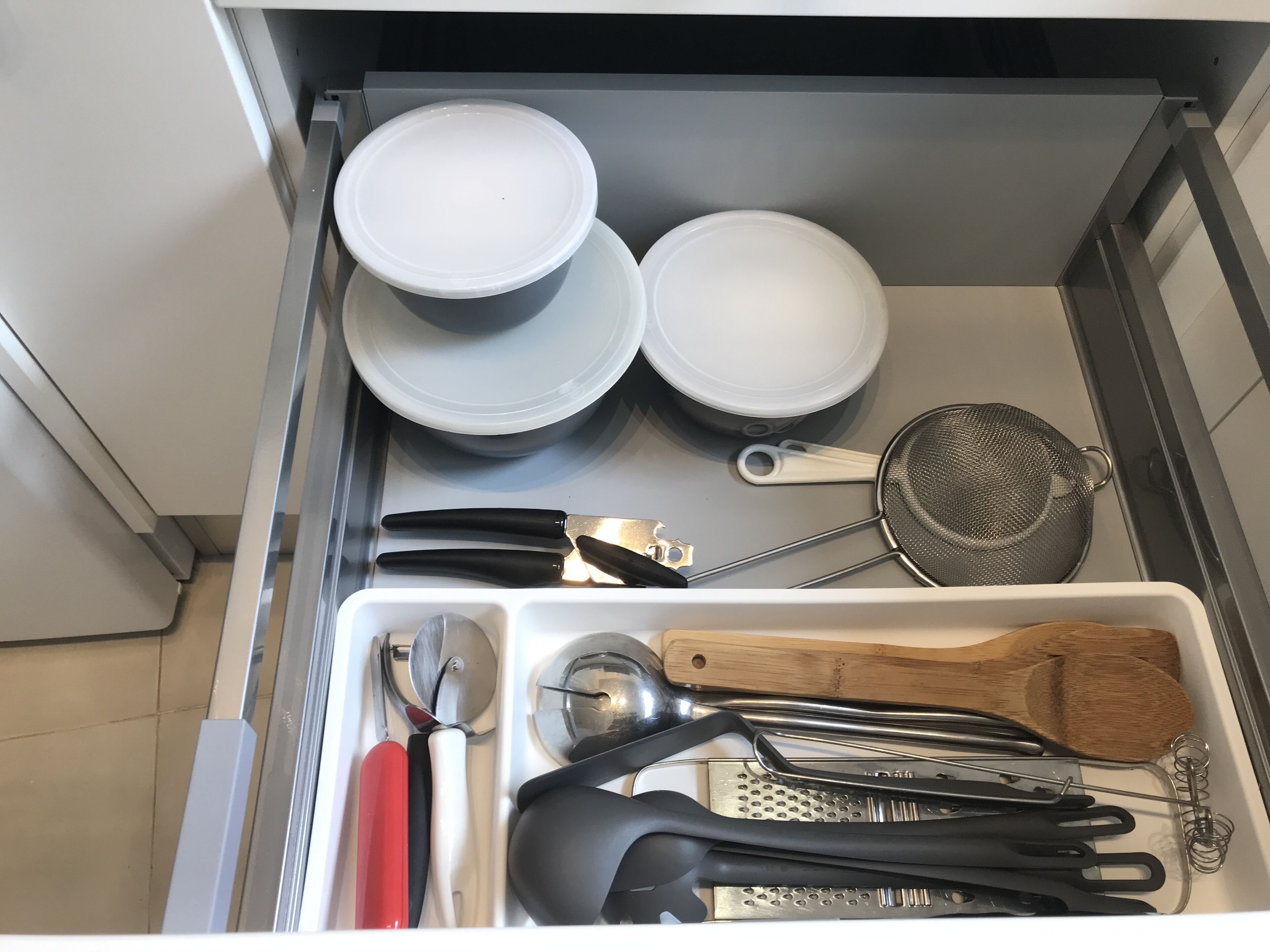 A kitchen drawer with various utensils, including a grater, tongs, and a strainer.
