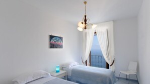 A bedroom with two beds, a nightstand, a lamp, a picture on the wall, and a view of the sea through a window with curtains. - Moressas two units accommodates up to 9 (Praiano)