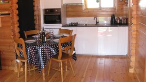 A wooden cabin interior with a dining table set for four, a kitchen with white cabinets, and a window above the sink.