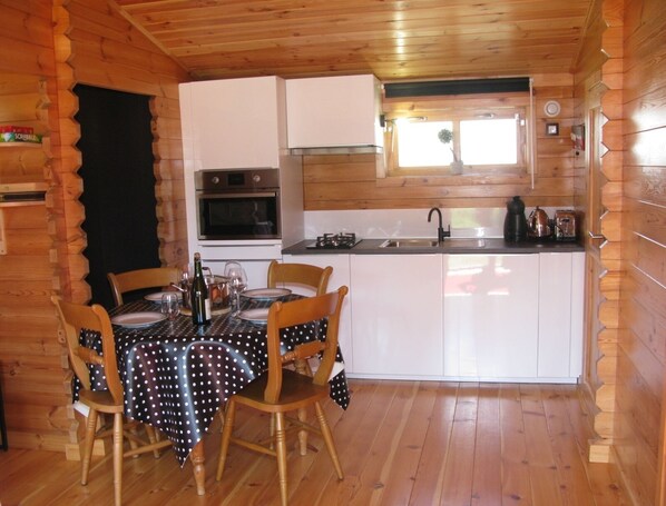 L’intérieur d’un chalet en bois comprend une salle à manger avec une table dressée pour quatre personnes, une cuisine avec des meubles blancs et une fenêtre au-dessus de l’évier.