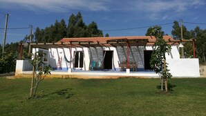 A white building with a red-tiled roof, a covered porch, and a small garden area with grass and a few trees. - "Casa da Charca" - SW Alentejano (Odemira)