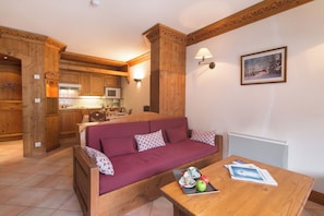 A cozy room with a wooden sofa, a dining table with a tray of food, and a kitchen area in the background.