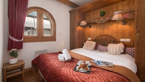 Une chambre confortable avec une tête de lit en bois, une grande fenêtre avec des rideaux, une table de chevet ornée d’une plante en pot et un lit bien fait, recouvert d’une couverture rouge.