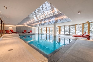 An indoor swimming pool with clear glass ceiling, red lounge chairs, and large windows.