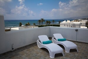 Un balcon avec deux fauteuils blancs, dont l’un est agrémenté d’un coussin turquoise, donnant sur une zone côtière bordée de palmiers et de bâtiments.