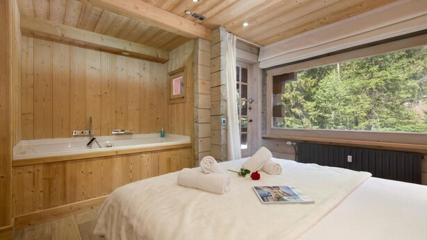 Ein holzgetäfeltes Zimmer im Chalet-Stil mit Holzboden, einem großen Fenster, einer Badewanne, einem Bett mit Handtüchern und einer Zeitschrift.