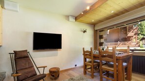 Ein Zimmer mit einem Holztisch, Stühlen, einem an der Wand montierten Fernseher und einem rustikalen Hocker.