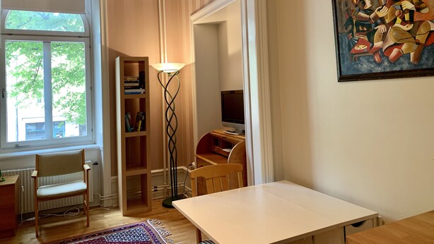 Ein Zimmer mit großem Fenster, Stehlampe, Gemälde, Schreibtisch und Stuhl.