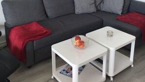 A modern living room with a grey sectional sofa, two white side tables, a red throw blanket, and a painting of beach chairs on the wall. - Apartment with a dream view directly on the Pearl Bay (Büsum)