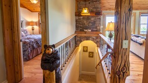 Un escalier en bois surmonté d’une sculpture d’ours, une cheminée en pierre et, dans la pièce adjacente, un lit recouvert d’une couverture à carreaux.