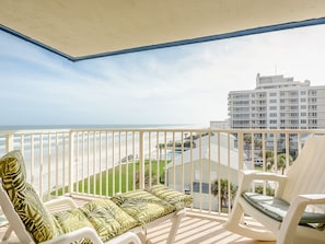 A balcony with white chairs and a sofa, overlooking a beach and a building. - 🏖️Top Floor Ocean View Retreat with Balcony and Pool, Close to Shopping and Dining | HA508🌊 (New Smyrna Beach)