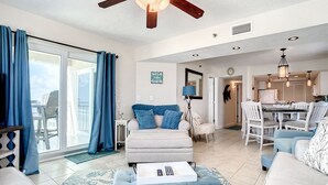 Ein geräumiger Wohnbereich mit Sofa, Hocker und einem Esstisch mit Stühlen. Ein Deckenventilator, ein großes Fenster mit Vorhängen und eine sichtbare Küche im Hintergrund.
