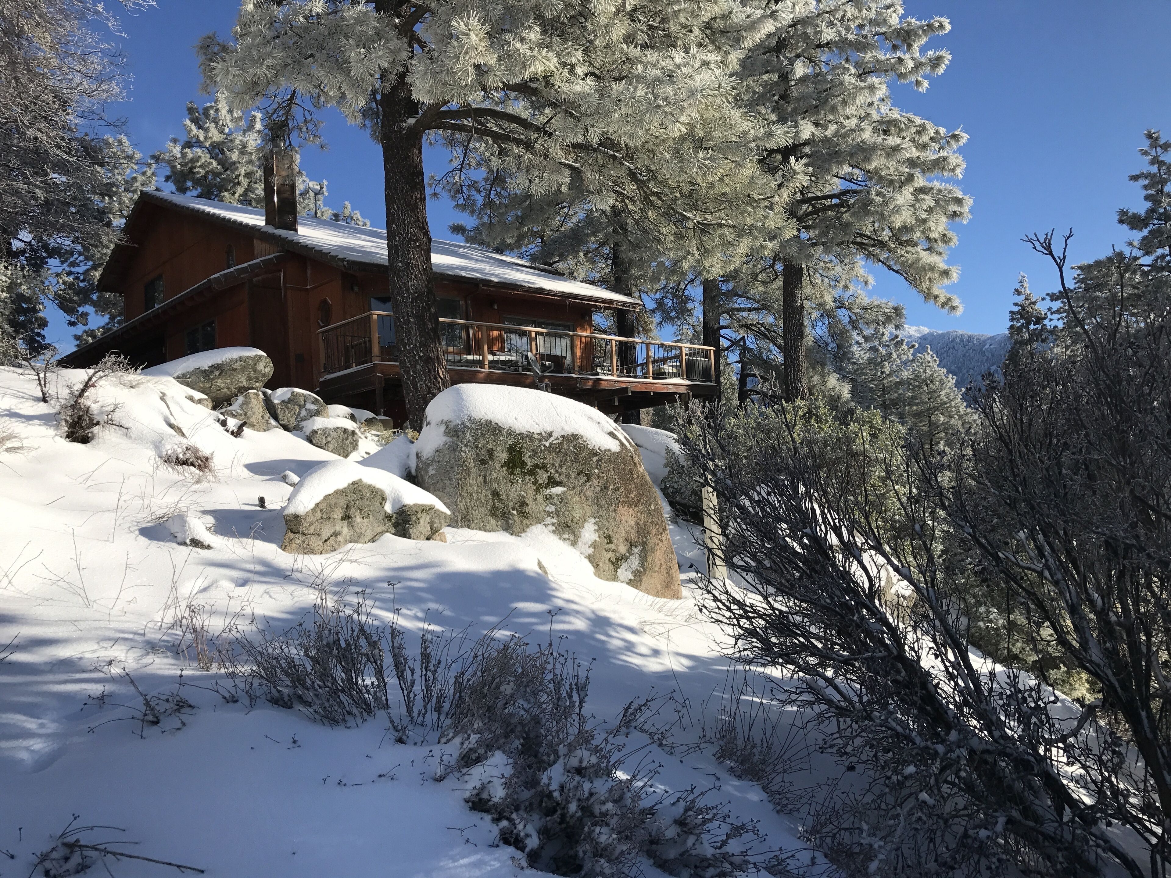 A wooden cabin with a balcony, surrounded by snow-covered trees and rocks.