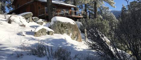 A wooden cabin with a balcony, surrounded by snow-covered trees and rocks.