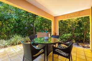 Un patio al aire libre con una mesa redonda de vidrio y sillas de mimbre, rodeado de exuberante follaje y un suelo de baldosas.