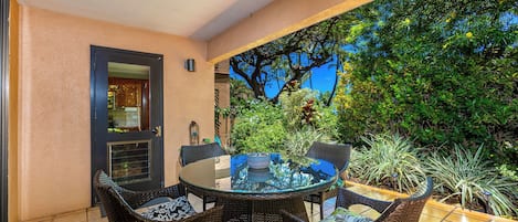 A covered patio with a round glass table and wicker chairs, surrounded by greenery and a view of trees.