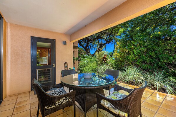 Un patio cubierto con una mesa redonda de vidrio y sillas de mimbre, rodeado de vegetación y vistas a árboles.