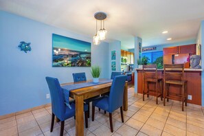 A dining area with a wooden table and blue chairs, a kitchen with wooden cabinets, and a view of the ocean through the window.