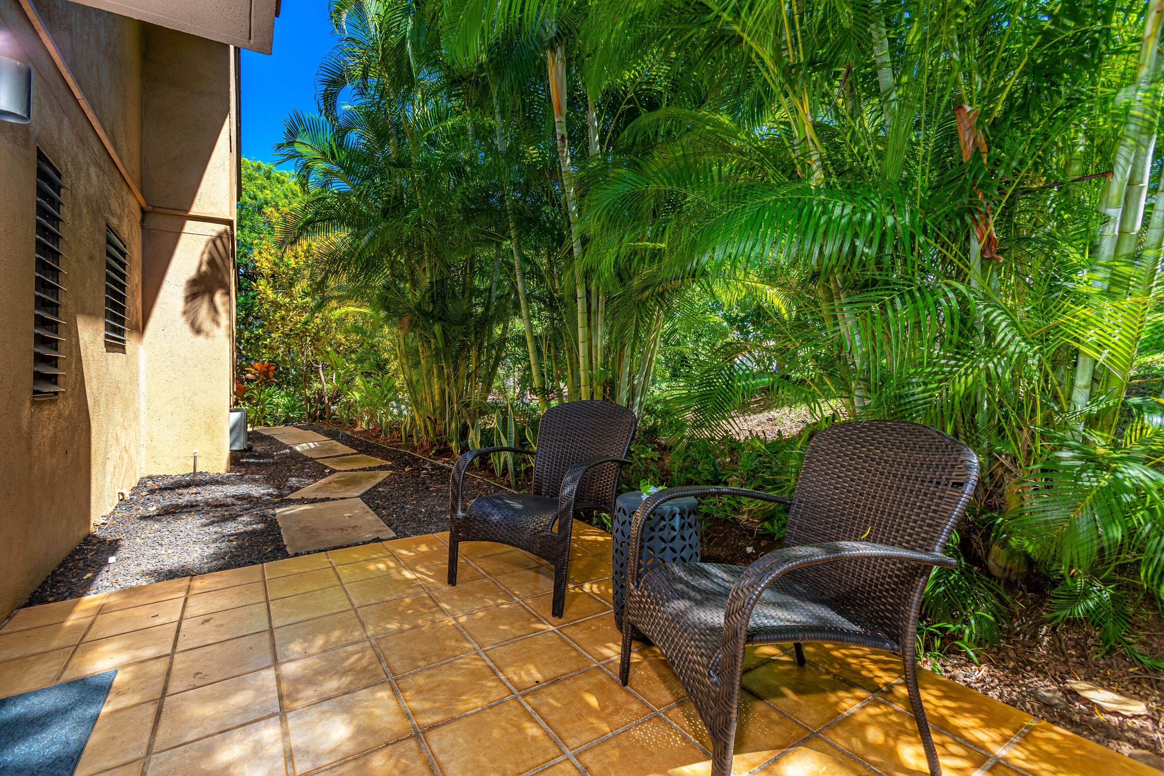 An outdoor patio with two wicker chairs, a tiled floor, and lush tropical vegetation.