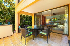 A glass-enclosed patio with a round table and wicker chairs, surrounded by potted plants and a view of the garden.