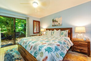 A bedroom with a large bed, a wooden nightstand, a ceiling fan, and a view of a tropical garden through a sliding glass door.