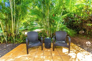 Two wicker chairs and a small table in a garden area with a tiled floor, surrounded by lush green foliage.