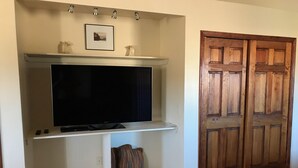 Une pièce avec un téléviseur fixé au mur, placé sur une étagère blanche, une petite photo encadrée au-dessus, et une porte en bois avec un motif en panneaux.