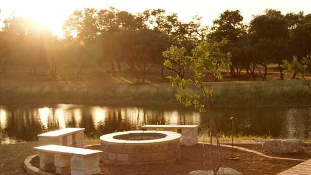 A circular stone fire pit surrounded by stone benches, set in a landscaped area with a pond and trees in the background. - Private 36-Acre Ranch Near Austin & Dripping Springs - Pond & Fire Pit (Driftwood)