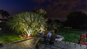 Een terras buiten met een tafel en stoelen, omgeven door een tuin met palmbomen en een hek.