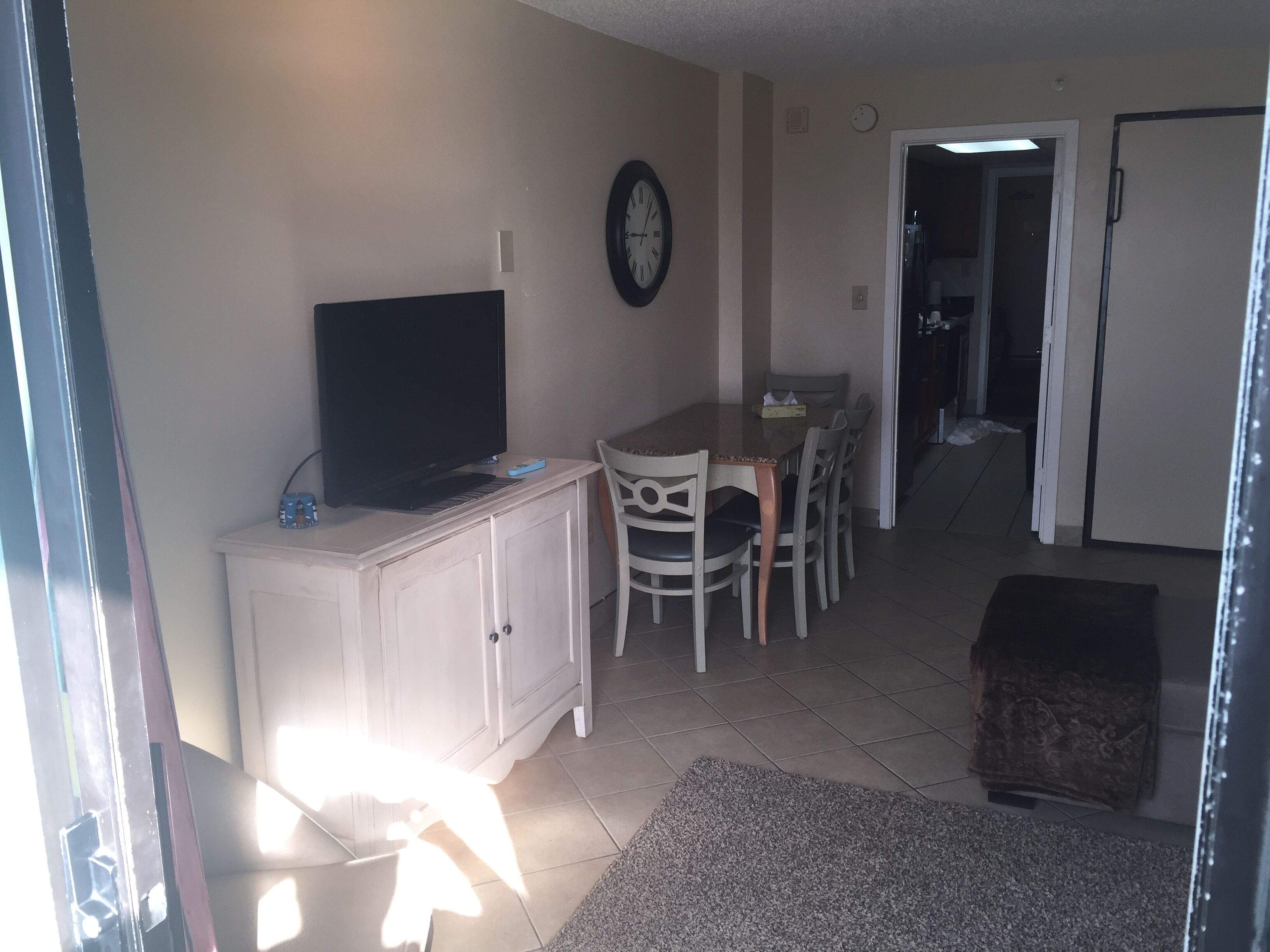 A living room with a TV on a cabinet, a dining table with chairs, a clock on the wall, and a door leading to another room.