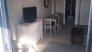 Una sala de estar con un televisor sobre un mueble, una mesa de comedor con sillas, un reloj en la pared y una puerta que da a otra habitación.
