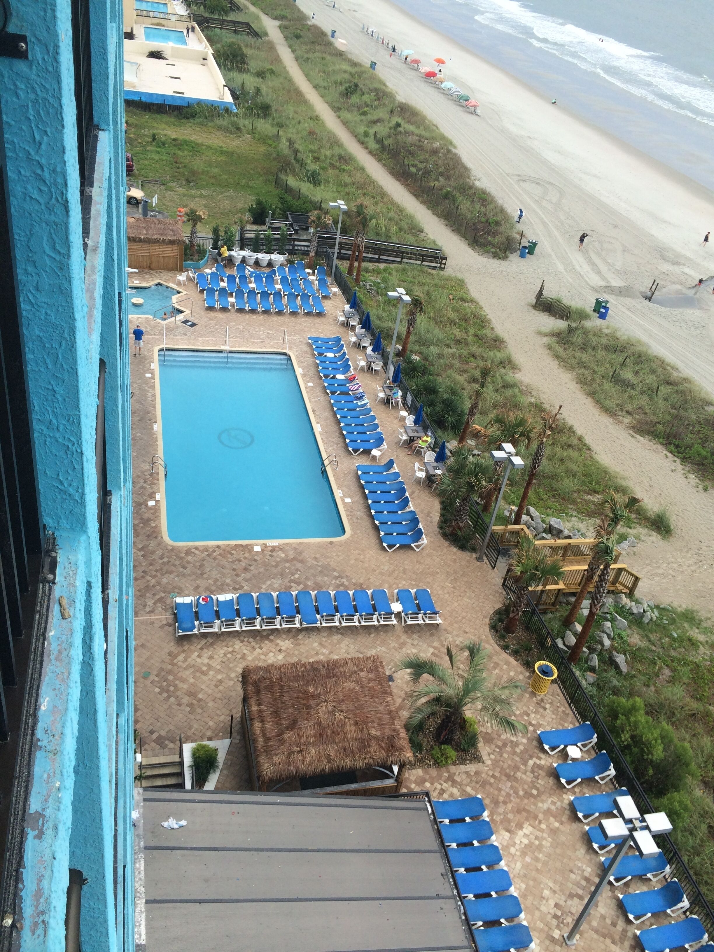 A pool area with blue chairs and a large pool, surrounded by a sandy beach and greenery.