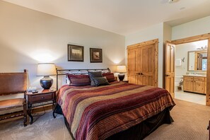 A bedroom with a large bed, wooden wardrobe, bedside tables with lamps, a chair, and framed pictures on the wall.
