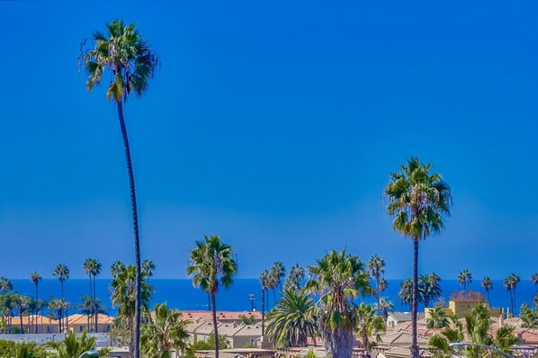 A coastal area with palm trees and buildings, likely a resort or hotel area with clear skies. - Just Blocks To Beach, Spanish Style Townhome With Panoramic Ocean View (Oceanside)