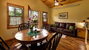 A dining area with a wooden table, chairs, and a vase of flowers. There is a brown leather couch and a small wooden cabinet with a lamp.