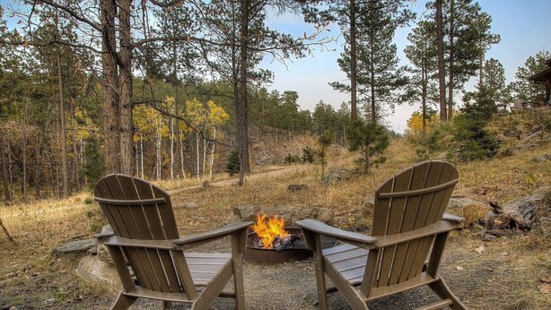 Two Adirondack chairs facing a campfire in a forested outdoor setting.