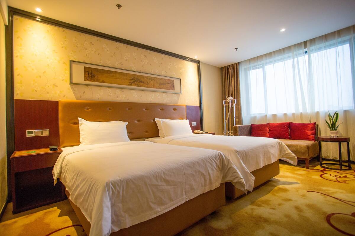 Habitación de hotel con dos camas, cabeceros, mesitas de noche, una lámpara, una ventana con cortinas y una planta.